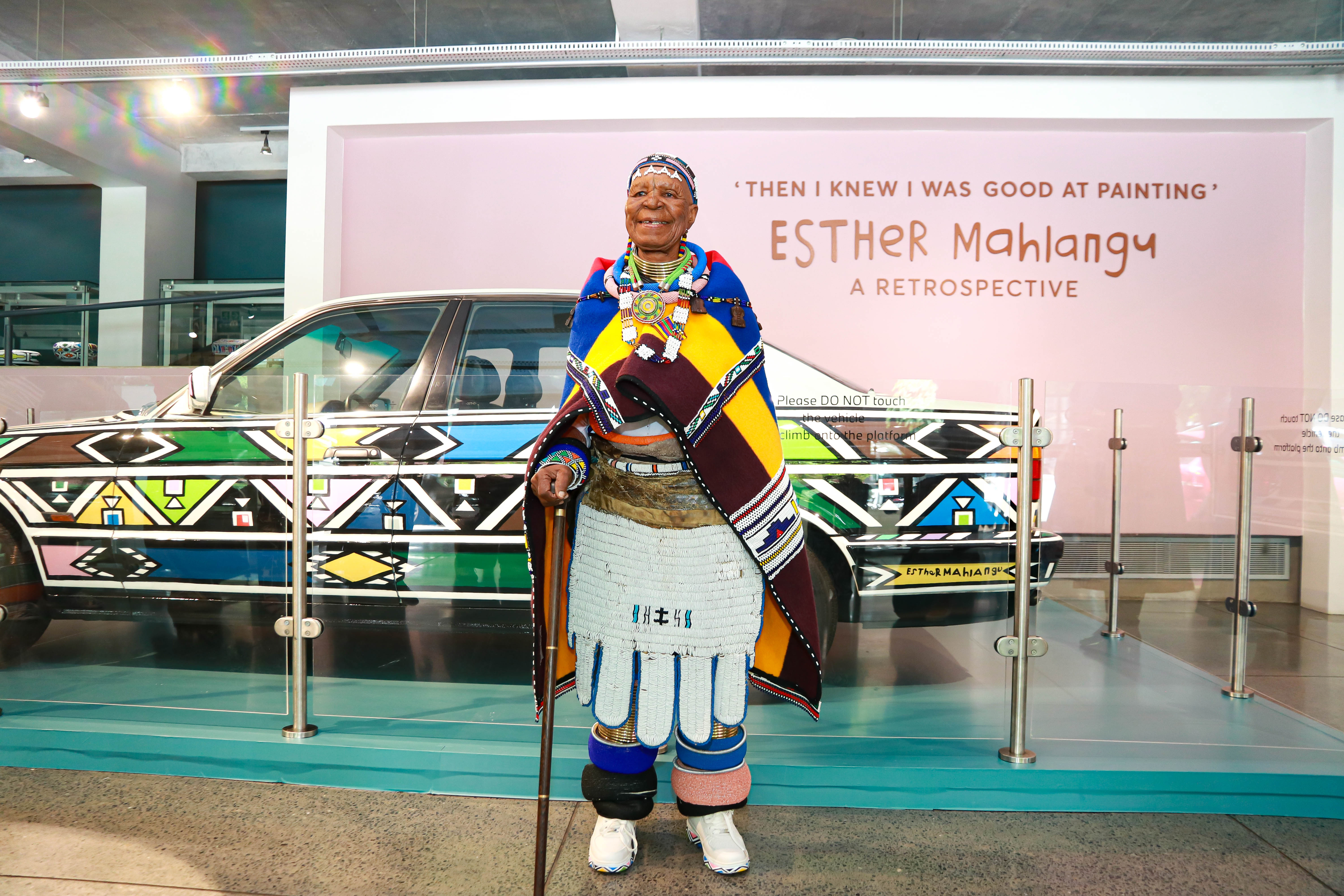 Dr Esther Mahlangu standing before her hand-painted BMW Art Car at the 'Then I Knew I Was Good at Painting' retrospective at Wits Art Museum