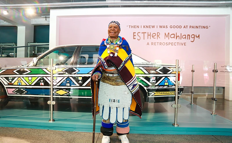 Dr Esther Mahlangu standing before her hand-painted BMW Art Car at the 'Then I Knew I Was Good at Painting' retrospective at Wits Art Museum