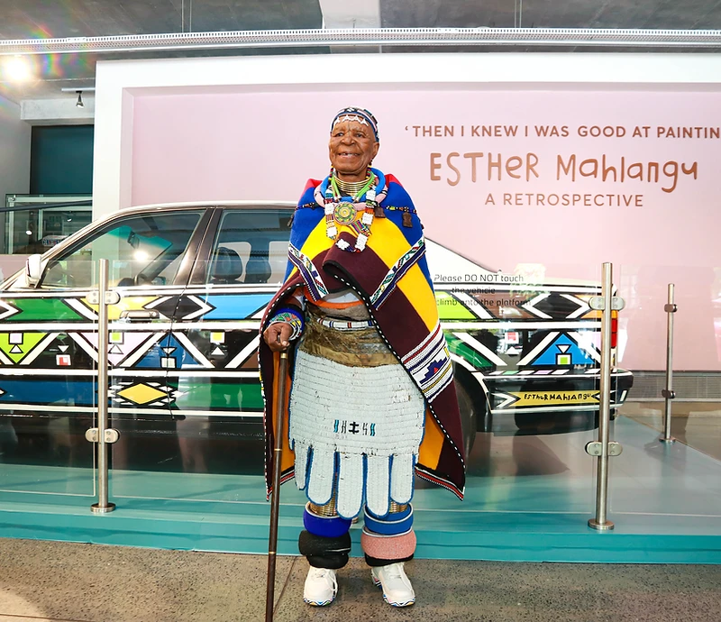 Dr Esther Mahlangu standing before her hand-painted BMW Art Car at the 'Then I Knew I Was Good at Painting' retrospective at Wits Art Museum