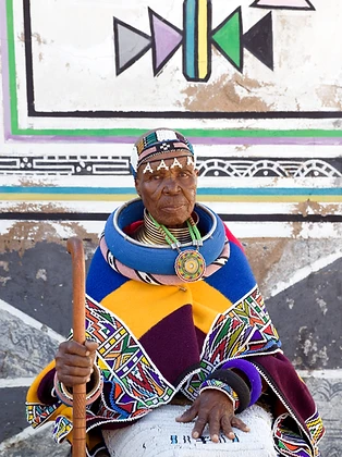 Seated portrait of Esther Mahlangu with a walking stick before a mural, by Clint Strydom