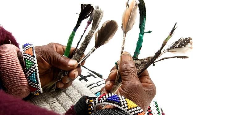 Hands holding handmade feather brushes with beaded bracelets — Dr Esther Mahlangu's first brushstrokes