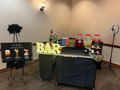 Signature drinks bar setup with custom cocktail menu easel, light-up BAR marquee, and beverage dispensers at a private event