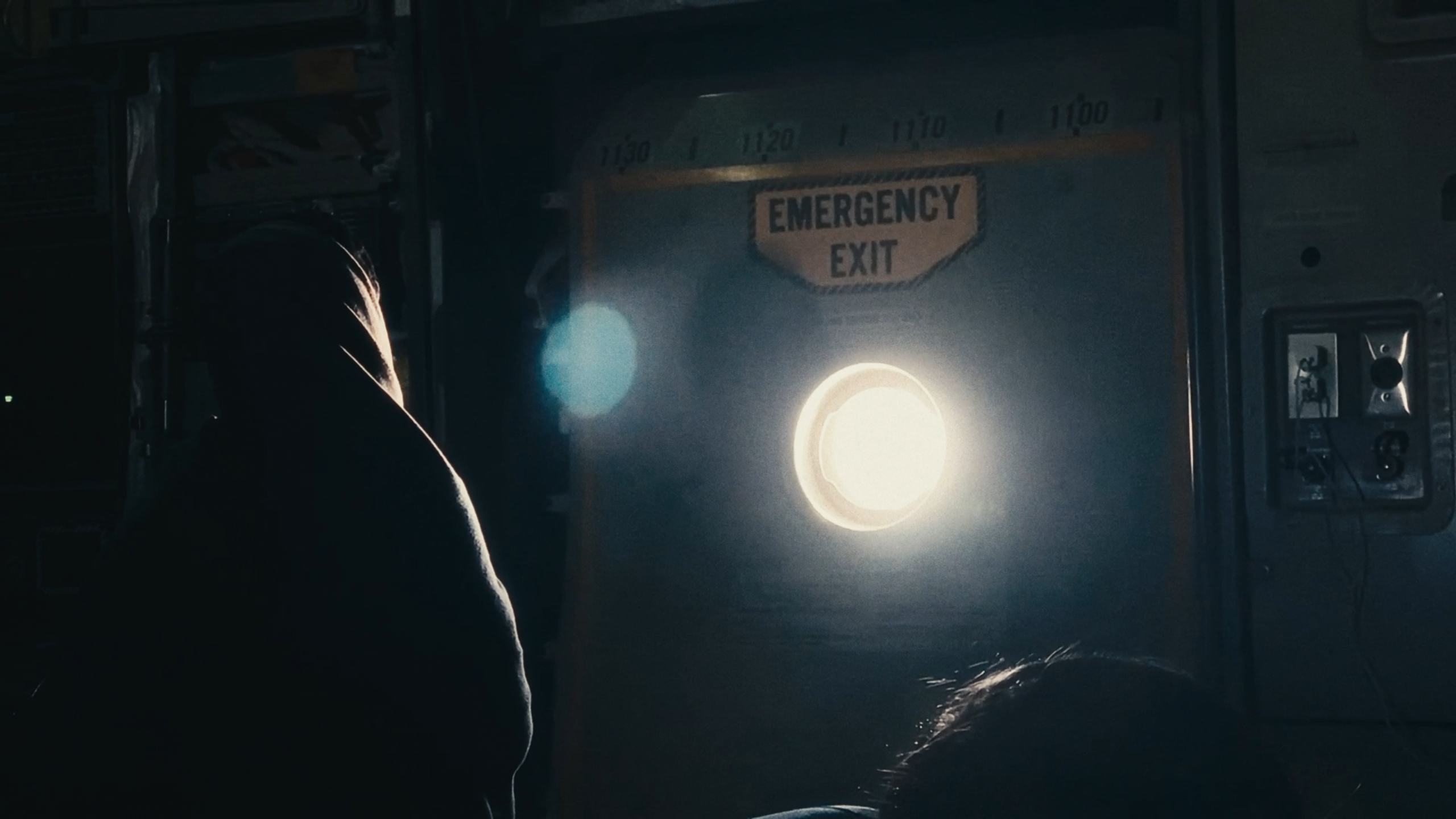 A hooded figure before an emergency exit door, lit by a porthole flare