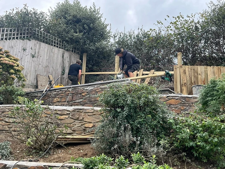 Two workers building a tiered stone retaining wall and timber fence
