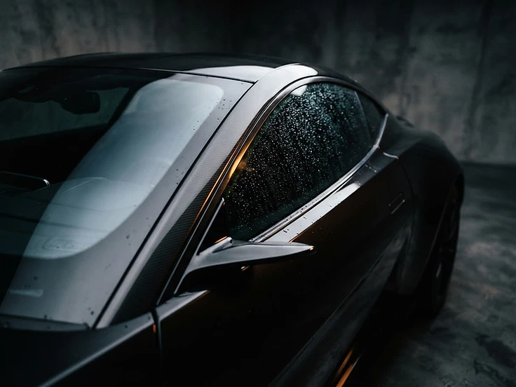 TOKYOFILMS Ceramic Tint poster — a dark metallic sports car in a minimalist concrete detailing studio under dramatic rim lighting, with a macro close-up of the side window showing deep ceramic tint with a cool silver-blue reflection and water beading on the glass.