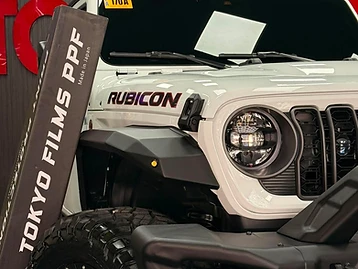 White Jeep Rubicon with aftermarket fenders parked in the TOKYOFILMS PPF installation bay beside branded signage.