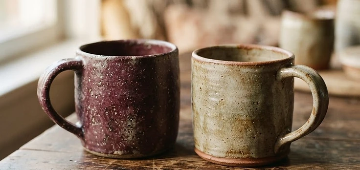 Two handmade ceramic mugs resting on a wooden table in warm window light
