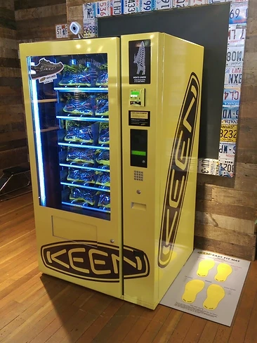 Bright yellow KEEN branded vending machine dispensing shoes in a wood-walled space