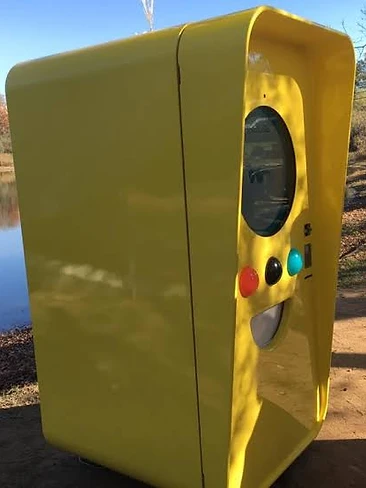 Bright yellow Snapbot kiosk with a circular display standing outdoors on a dirt path by a pond