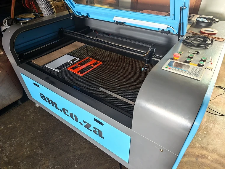 KEVMAR's CO₂ laser cutting and engraving machine in operation — lid open, honeycomb cutting bed visible with an orange laser-cut component on the bed.