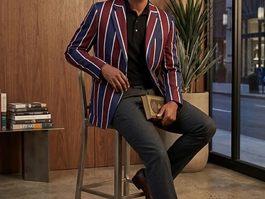 A model wearing a bespoke burgundy, navy and white striped sports blazer, seated in a modern interior — illustrating the Club Blazers group ordering process.