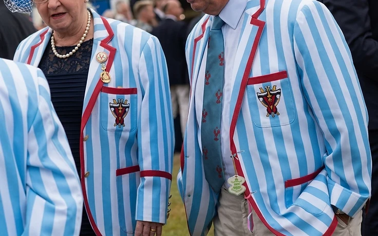 Two members wearing matching sky-blue and white striped bowls blazers with red piping and an embroidered gold chalice crest at an outdoor bowls event in Australia.