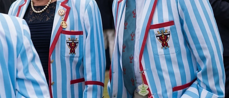 Two members wearing matching sky-blue and white striped bowls blazers with red piping and an embroidered gold chalice crest at an outdoor Australian bowls event.