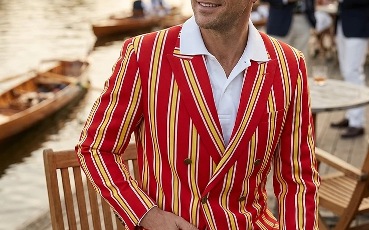 Man wearing a double-breasted rowing blazer with red, yellow, and white stripes at a riverside table during golden hour, with other rowing club members in the background