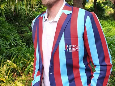 A man wearing a bespoke Help for Heroes Cricket Team blazer with light blue, navy, and terracotta vertical stripes and an embroidered Help for Heroes crest, standing in a tropical garden in bright sunlight.
