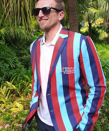 A man wearing a bespoke Help for Heroes Cricket Team blazer with light blue, navy, and terracotta vertical stripes and an embroidered Help for Heroes crest, standing in a tropical garden in bright sunlight.