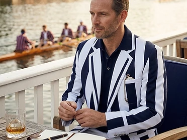 Man wearing a navy and white striped rowing blazer seated at a riverside terrace overlooking a regatta scene with a rowing crew on the water