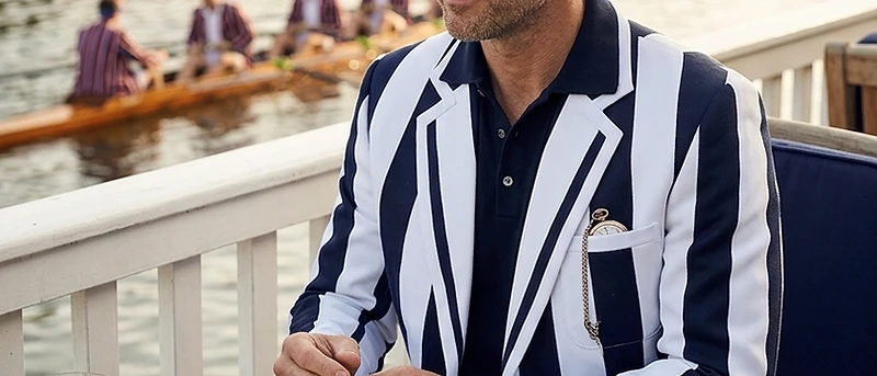 Man wearing a navy and white striped rowing blazer seated at a riverside terrace overlooking a regatta scene with a rowing crew on the water