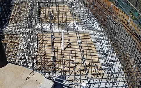 High-angle view of a rectangular footing reinforcing mat set inside plywood formwork, with vertical PVC utility penetrations rising from the subgrade