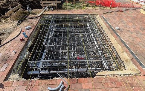 High-angle view into a rectangular excavated fountain pit with a dense #5 rebar grid, white PVC plumbing, and yellow string lines over red brick pavers