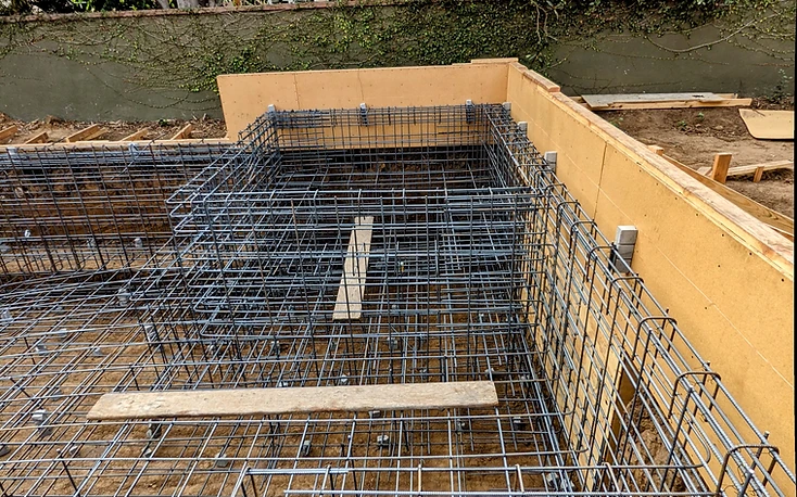 Rebar cage standing against wooden formwork with planks laid across the top of a pool shell