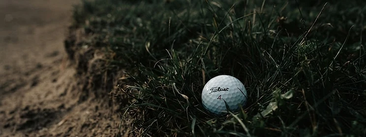 A golf ball resting in dark rough grass beside a bunker.