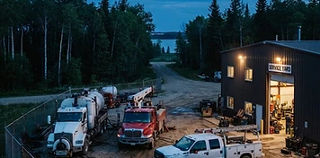 Oilfield service yard with crew trucks, vacuum truck and picker at a fenced shop near Bonnyville at dusk