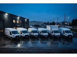 Row of white cargo vans and a box truck in a commercial service yard at blue-hour dusk