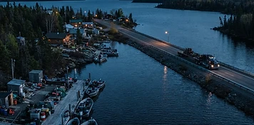 Northern Saskatchewan community on a channel between two lakes, work boats at a dock and a tractor-trailer crossing the causeway at dusk