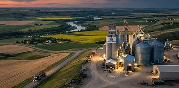 Seed plant and grain facility above river-valley farmland with a grain truck on a valley road near Peace River at dusk