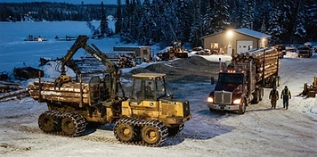 Forestry service yard with log truck and forwarder under floodlights at a frozen lake edge in Lac La Biche at dusk