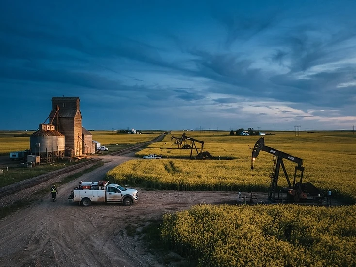 Oil pumpjacks in a golden canola field beside a country grain elevator at dusk in southeast Saskatchewan