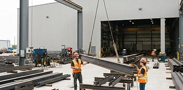 Steel fabrication crew guiding a lifted beam in a bright Manitoba fabrication yard