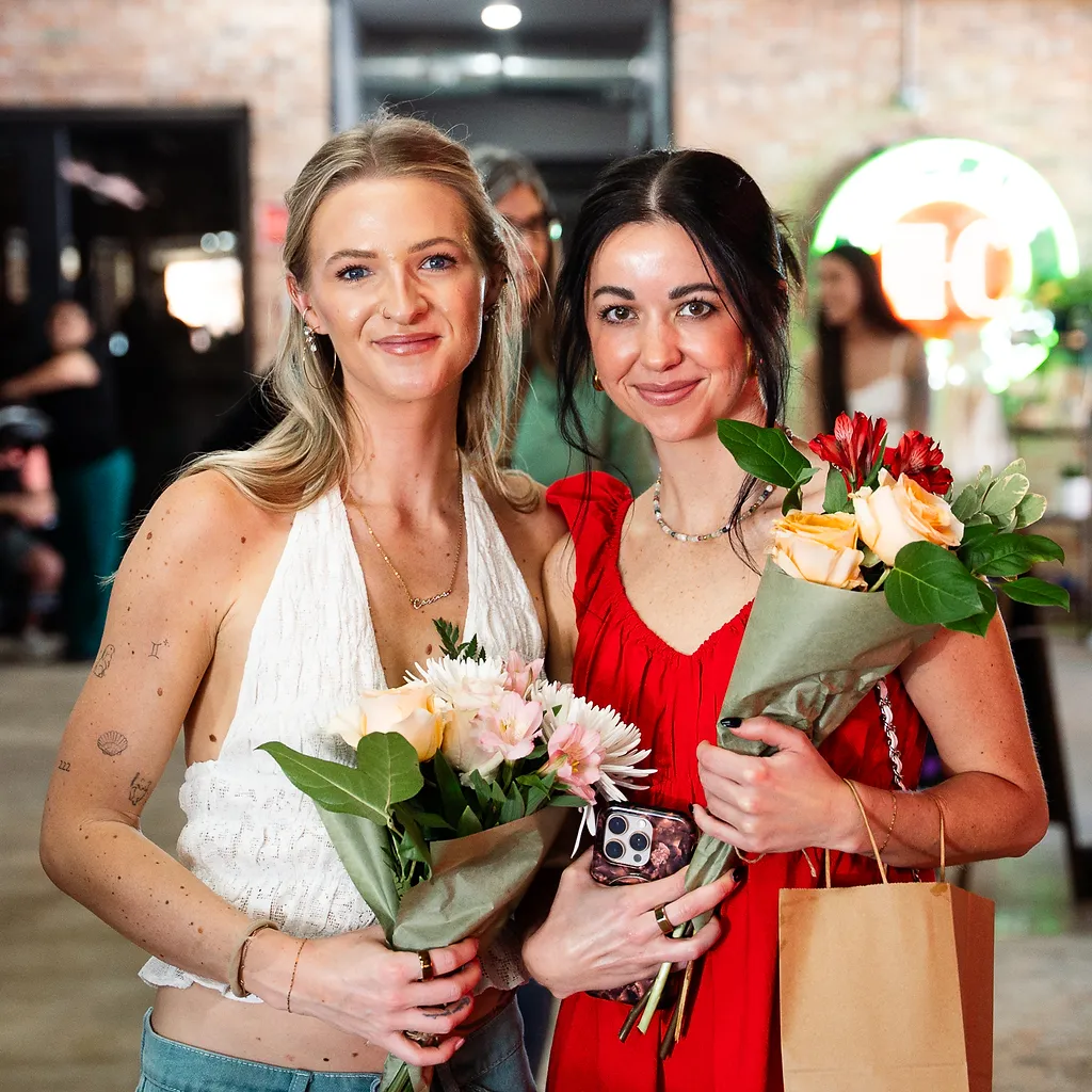 Two friends laughing together with fresh flower bouquets and a shopping tote at the market