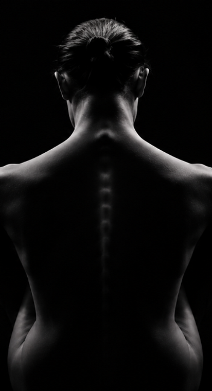 Black-and-white photograph of a person's bare back, head bowed, lit from behind