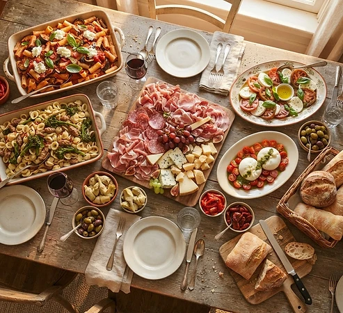 Overhead view of an Italian catering spread with pasta trays, cold cuts, bread, olives and salads on a rustic table