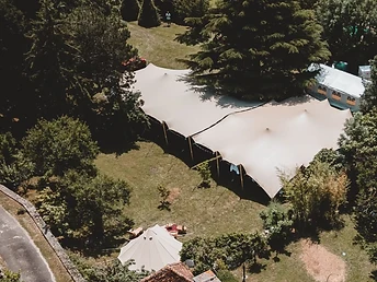 Vue aérienne d'une tente stretch beige dressée dans un parc arboré avec toiture en pierre