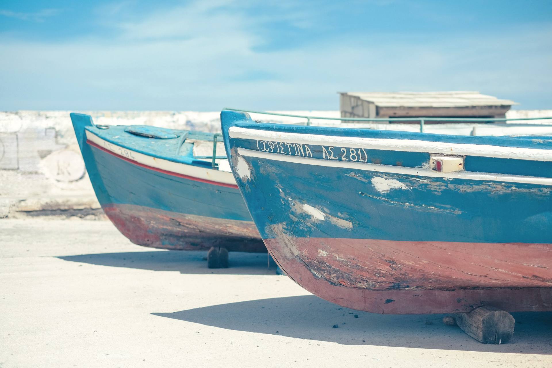 Two weathered wooden fishing boats resting on a pale shore, painted in faded blue and terracotta, under soft light.