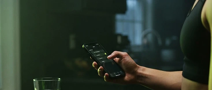 Mão marcando um hábito no celular ao amanhecer, ao lado de um copo de água e uma balança