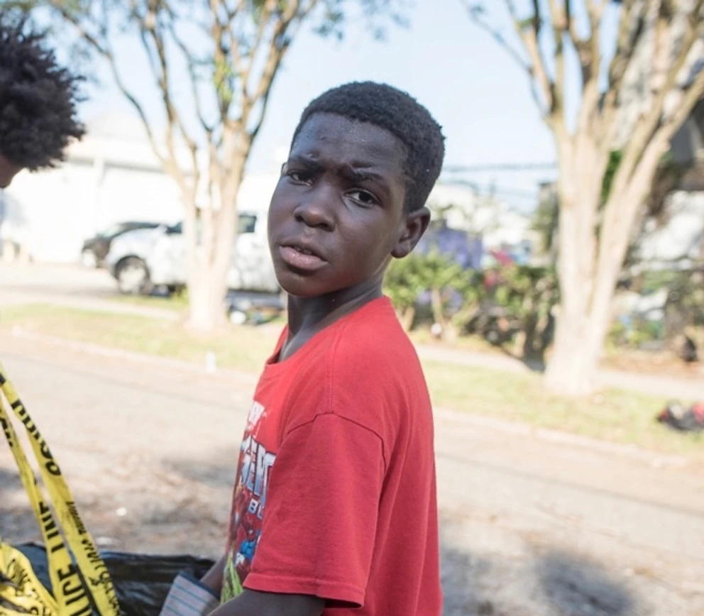 A young boy in a red Spider-Man shirt meets the camera on a sunlit North Baton Rouge street