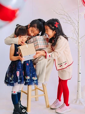 Three girls in holiday outfits holding wrapped gifts beside a lit white tree