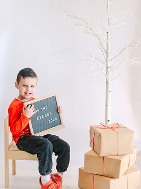 Boy on a wooden chair holding a Tis the Season 2026 letter board beside kraft paper gifts