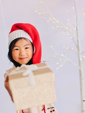 Girl in a Santa hat holding a gold wrapped gift out toward the camera