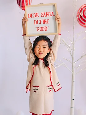 Girl holding a wooden Dear Santa, define good sign in front of a lit birch tree