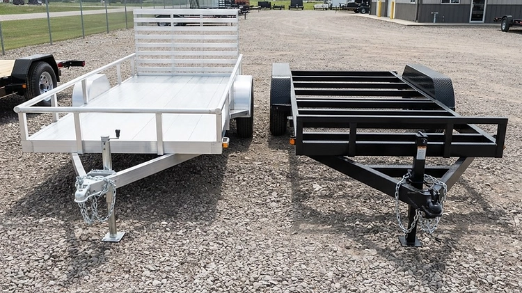 Side-by-side comparison of an extruded aluminum utility trailer and a heavy-duty painted steel equipment trailer parked on gravel