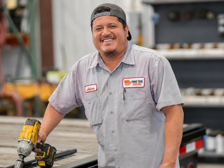 Prime Trail Trailers technician servicing a flatbed trailer with an impact wrench in the service bay