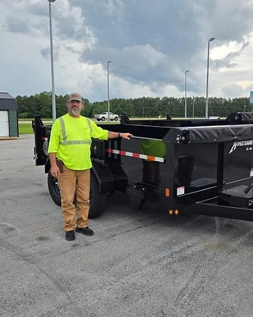 Thomas Barron, Prime Trail Trailers sales and support agent in a high-visibility shirt beside a black dump trailer at the dealership lot