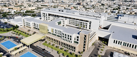 Aerial view of Sultan Qaboos Hospital in Salalah with white wings, palm-lined lawns, blue pools and the city skyline beyond