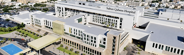 Aerial view of Sultan Qaboos Hospital in Salalah with white wings, palm-lined lawns, blue pools and the city skyline beyond