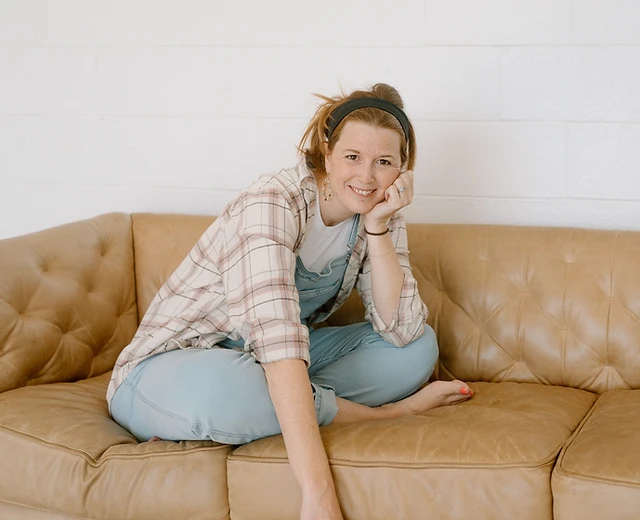 Katie Burke, motherhood photographer, seated on a tan leather sofa